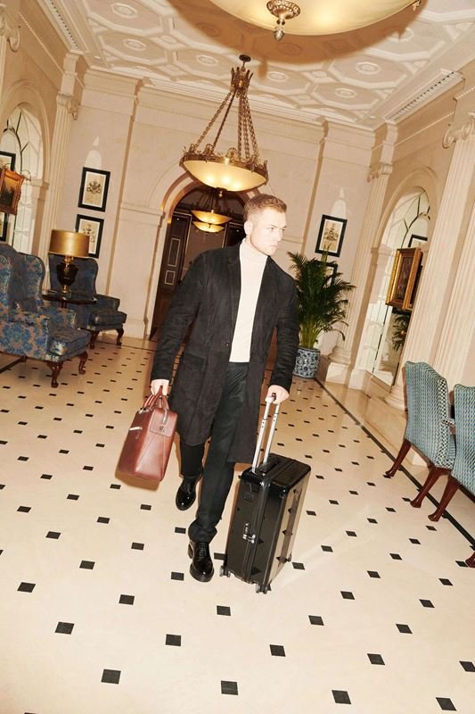 A man walks through a grand hotel lobby pulling a rolling suitcase and carrying a leather duffel bag, surrounded by elegant decor.
