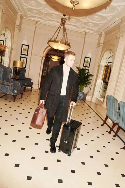 A man walks through a grand hotel lobby pulling a rolling suitcase and carrying a leather duffel bag, surrounded by elegant decor.