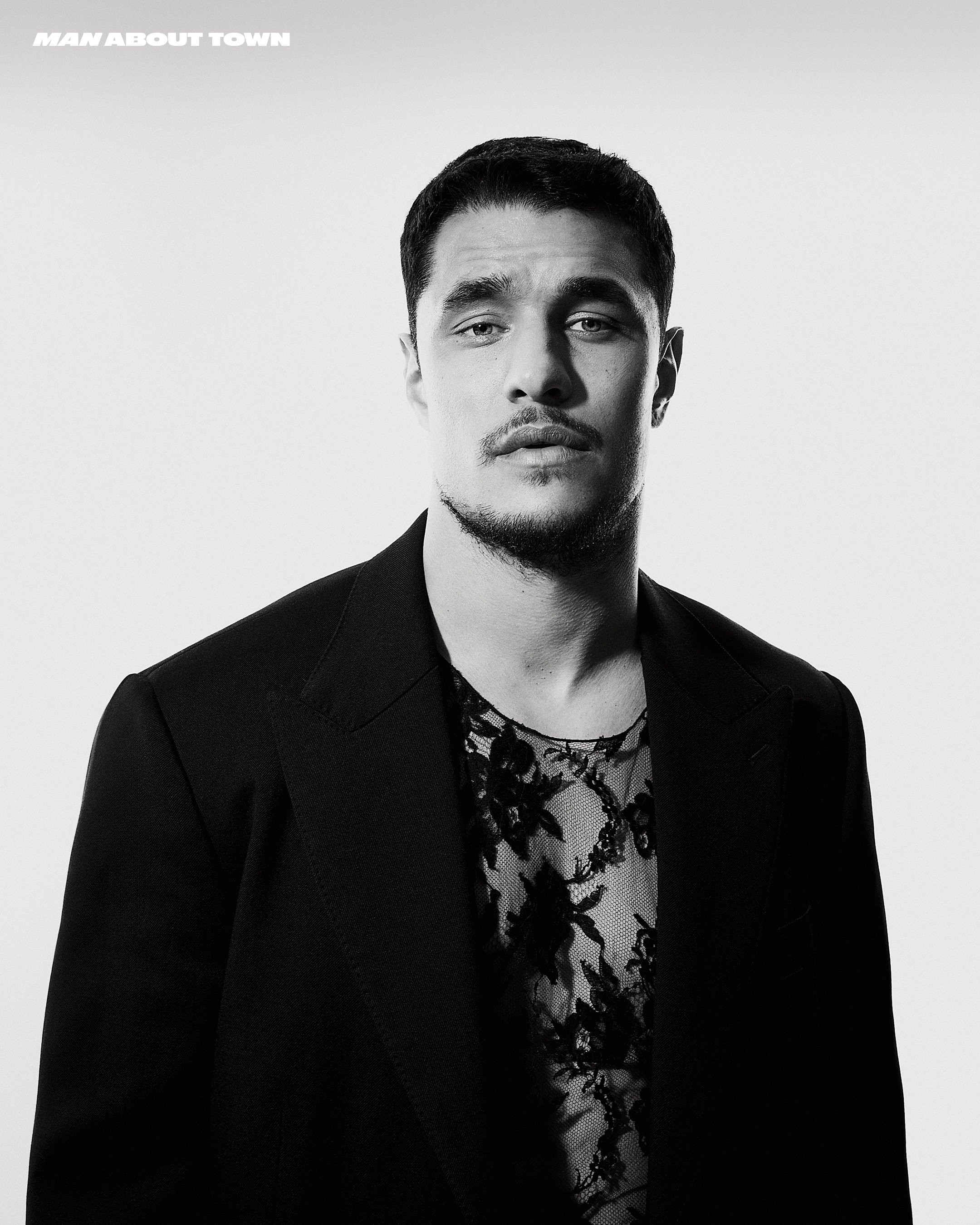 Man wearing a dark blazer over a sheer patterned shirt against a plain white studio background.