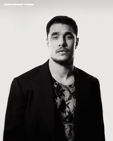 Man wearing a dark blazer over a sheer patterned shirt against a plain white studio background.