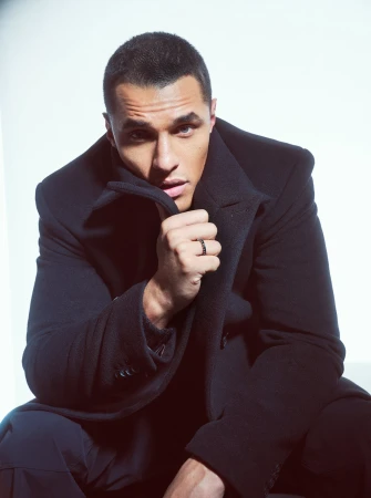 Man wearing a dark coat pulls up his collar, posing against a bright white background with hair styled by Charley Mcewen.