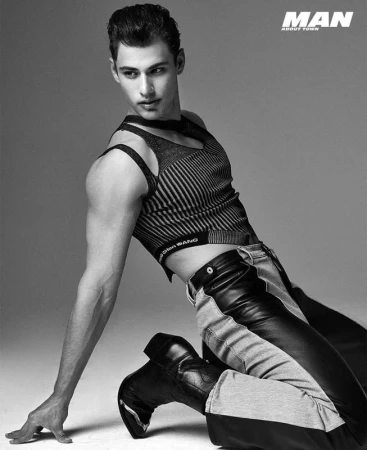 Monochrome fashion portrait of a male model kneeling in a striped crop top and leather trousers against a grey backdrop.