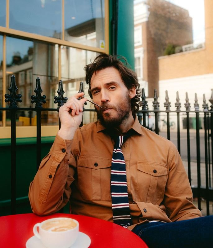 Man in brown shirt and striped tie sits at a red table with a coffee cup in an outdoor cafe with black wrought iron fence.