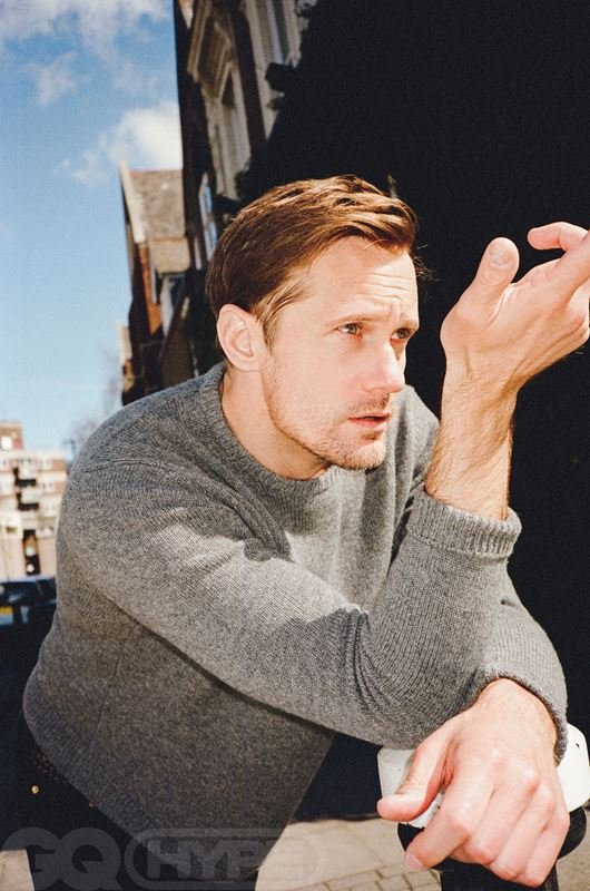 Portrait of Alexander Skarsgård in a grey sweater outdoors, with buildings and blue sky in the background, captured from a low angle.