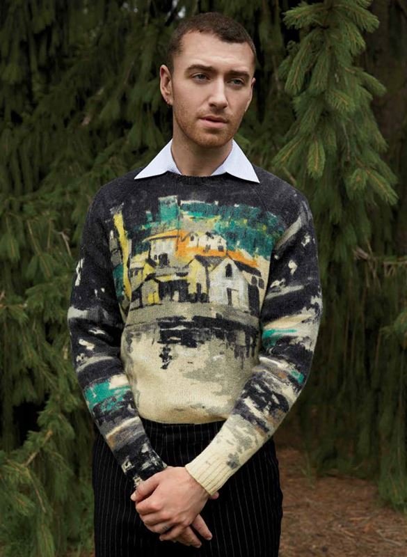 Portrait of a fair-skinned adult male wearing a sweater with a graphic design of houses and landscape, standing outdoors in front of green plants.