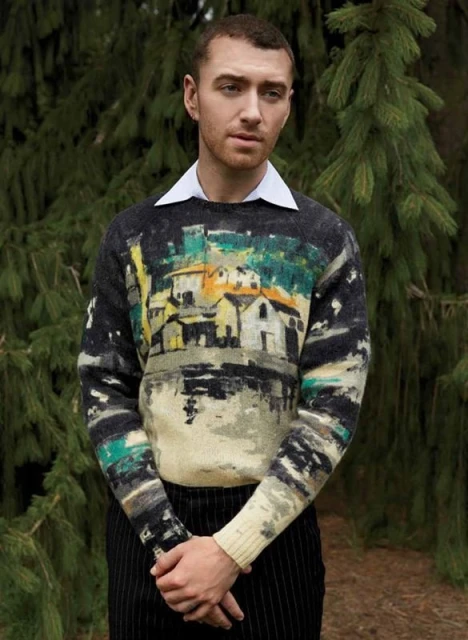Portrait of a fair-skinned adult male wearing a sweater with a graphic design of houses and landscape, standing outdoors in front of green plants.