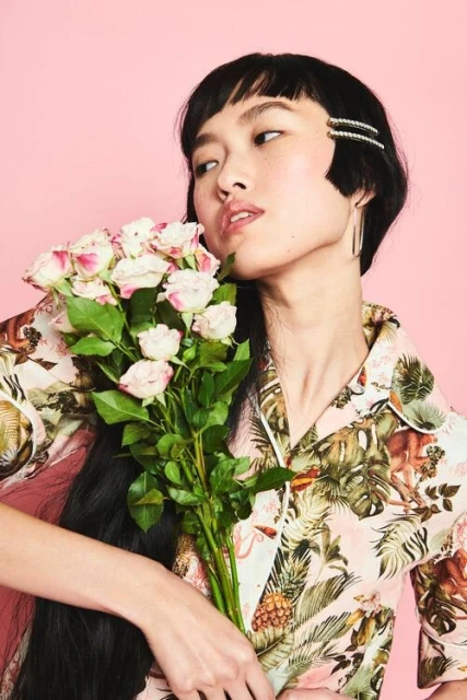 Asian woman in a floral shirt holds a bouquet of pink roses against a pink backdrop, creating a vibrant and eye-catching portrait.