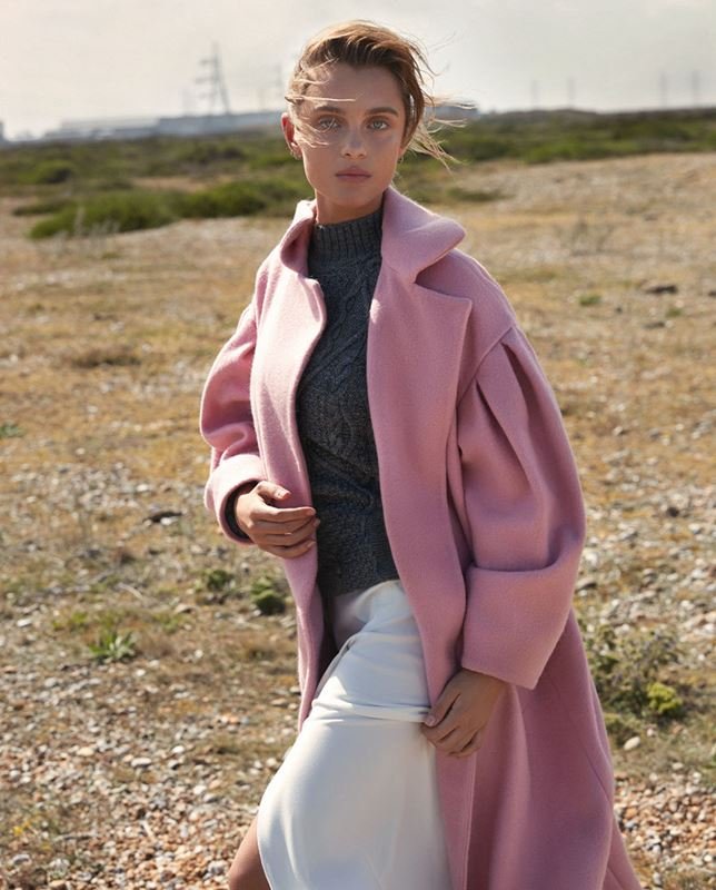 A fair-skinned adult female model poses outdoors in a pale pink coat, dark grey knit top, and white skirt on a sunny day.