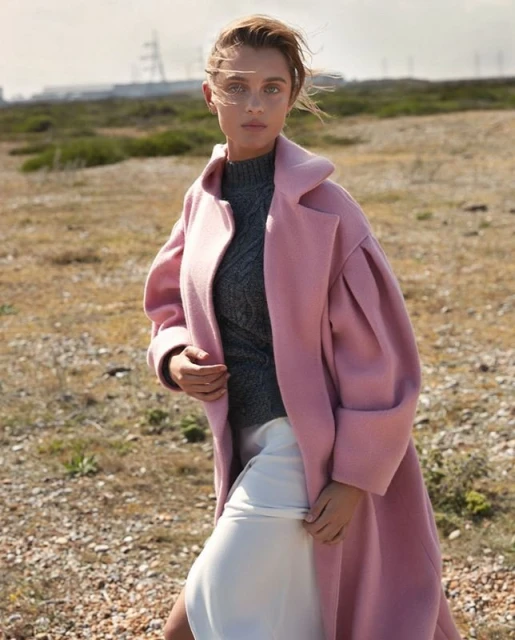 A fair-skinned adult female model poses outdoors in a pale pink coat, dark grey knit top, and white skirt on a sunny day.