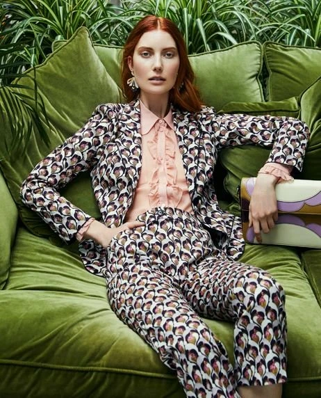 A red-haired woman poses on a green velvet couch in a patterned suit, holding a clutch. The background features green leafy plants.