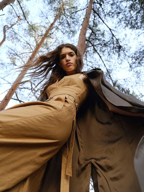 Low-angle shot of a woman in a tan jumpsuit and dark cloak in a forest with trees reaching toward a blue sky.
