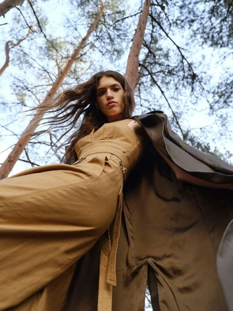 Low-angle shot of a woman in a tan jumpsuit and dark cloak in a forest with trees reaching toward a blue sky.