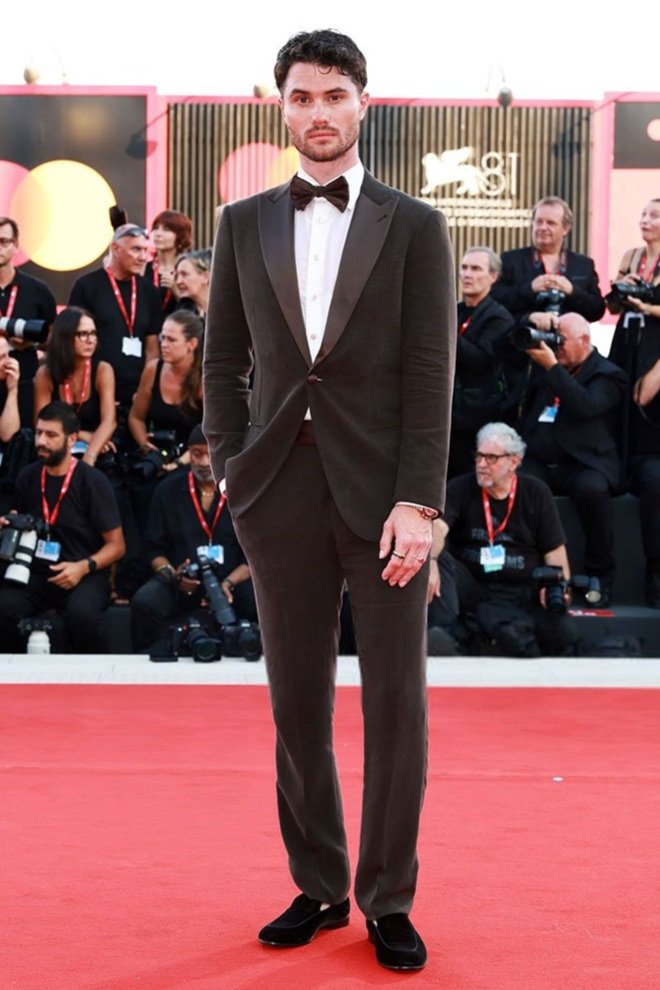 A full shot captures a dark-haired man in a brown suit with a bowtie on a red carpet, surrounded by people and camera equipment.