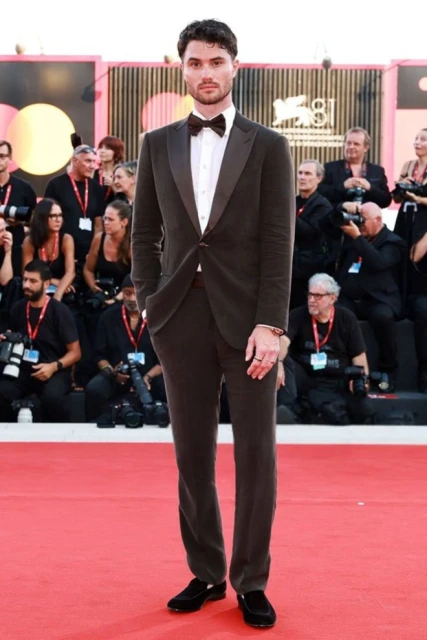 A full shot captures a dark-haired man in a brown suit with a bowtie on a red carpet, surrounded by people and camera equipment.