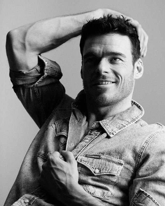 Black and white portrait of an adult male with hand on his head, wearing a denim shirt, indoors against a plain backdrop.