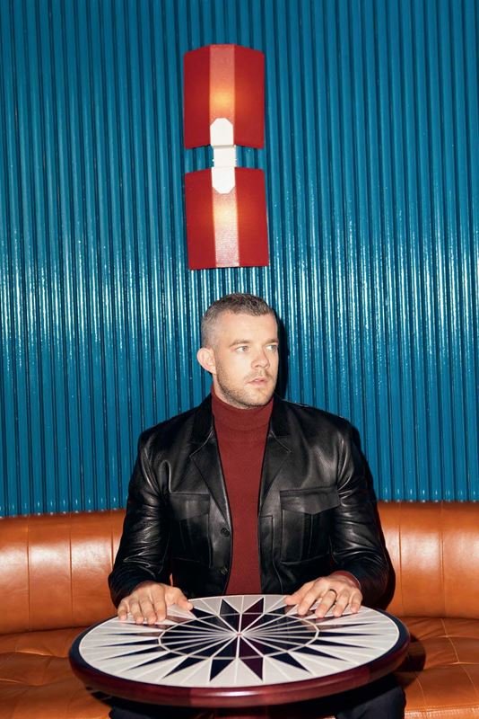 Portrait of a seated adult male with short hair and a dark jacket in an indoor setting with geometric patterned table and orange couch.
