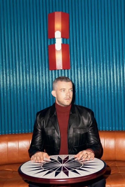 Portrait of a seated adult male with short hair and a dark jacket in an indoor setting with geometric patterned table and orange couch.