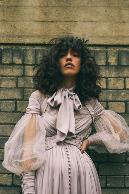Woman in a lavender gown with puffy sleeves and a bow poses against a brick wall, showcasing fashion and style in a portrait.