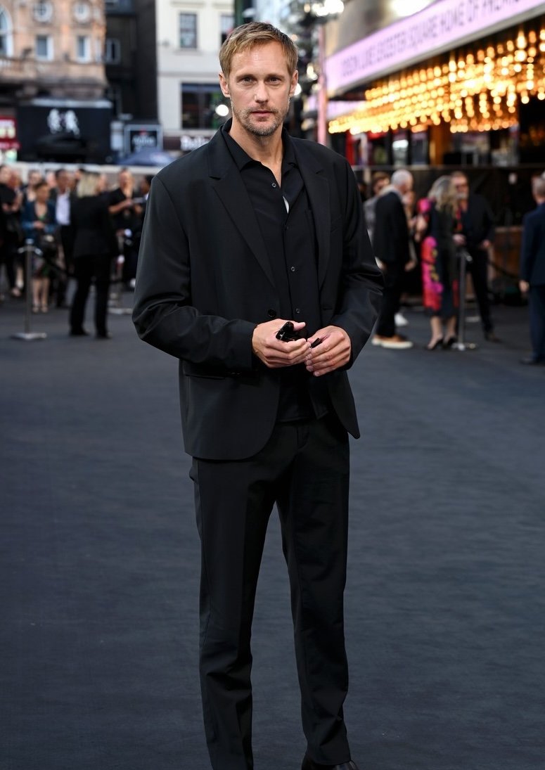 Alexander Skarsgård on the red carpet wearing a black suit and black shirt, posing for a portrait, in front of a theater.
