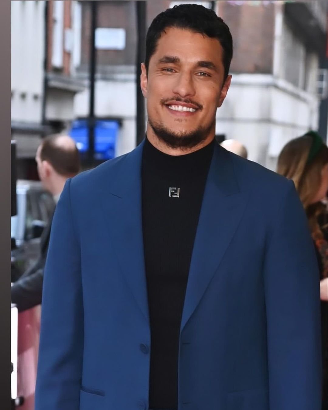 Smiling adult male in a blue blazer and black turtleneck, captured in a medium shot against an urban background with soft lighting.