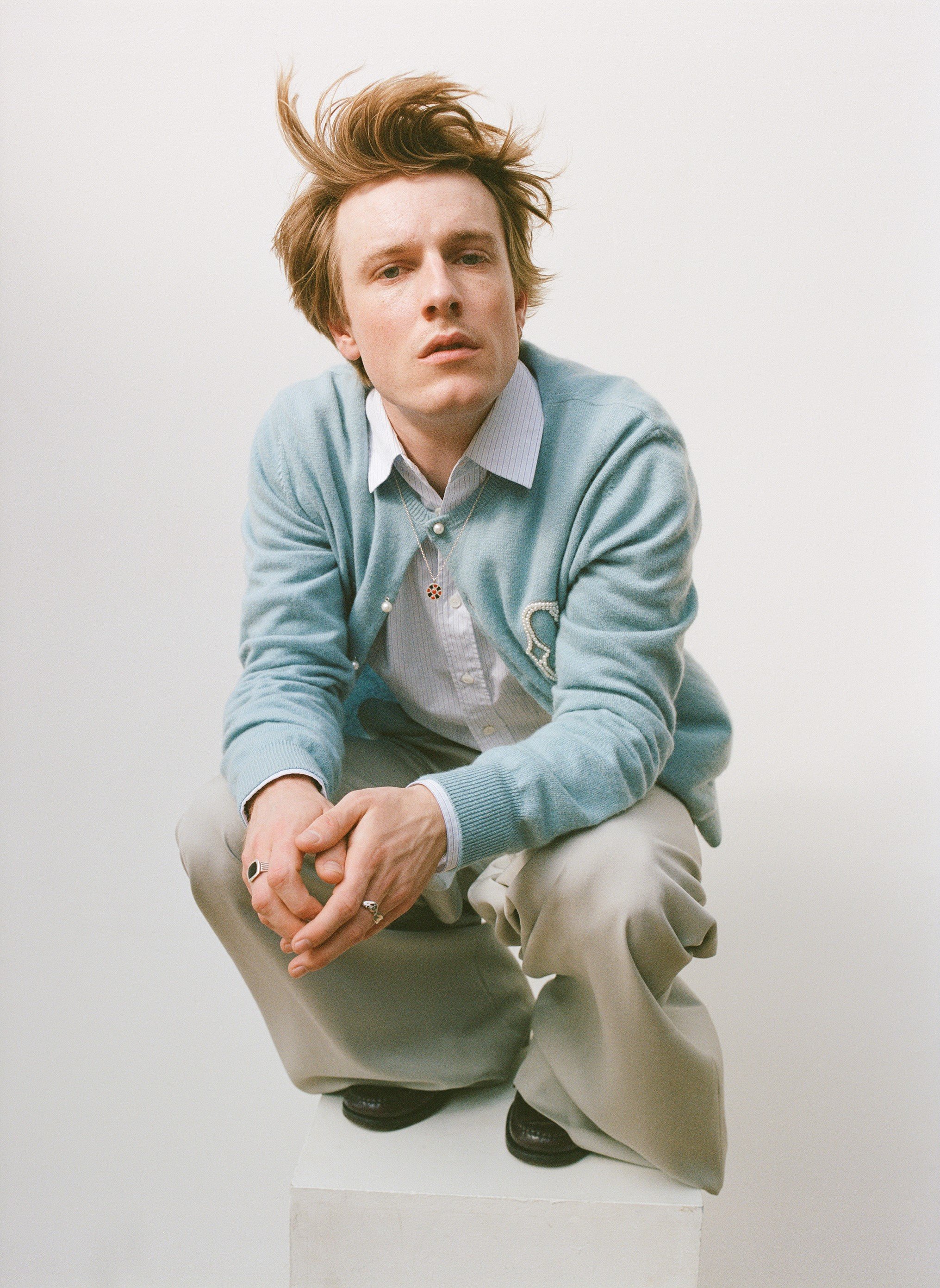 A young adult with reddish hair squats indoors, wearing a light blue cardigan, striped shirt, and tan pants, set against a white backdrop.