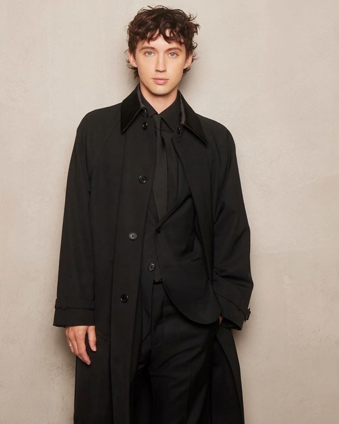 Portrait of adult male with curly brown hair wearing a black suit and overcoat indoors against a light beige wall.