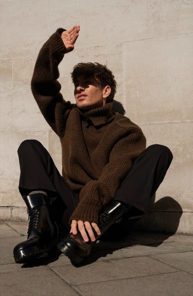 A young adult male poses outside wearing a brown knit sweater, dark trousers, and glossy black platform boots while shielding his face.
