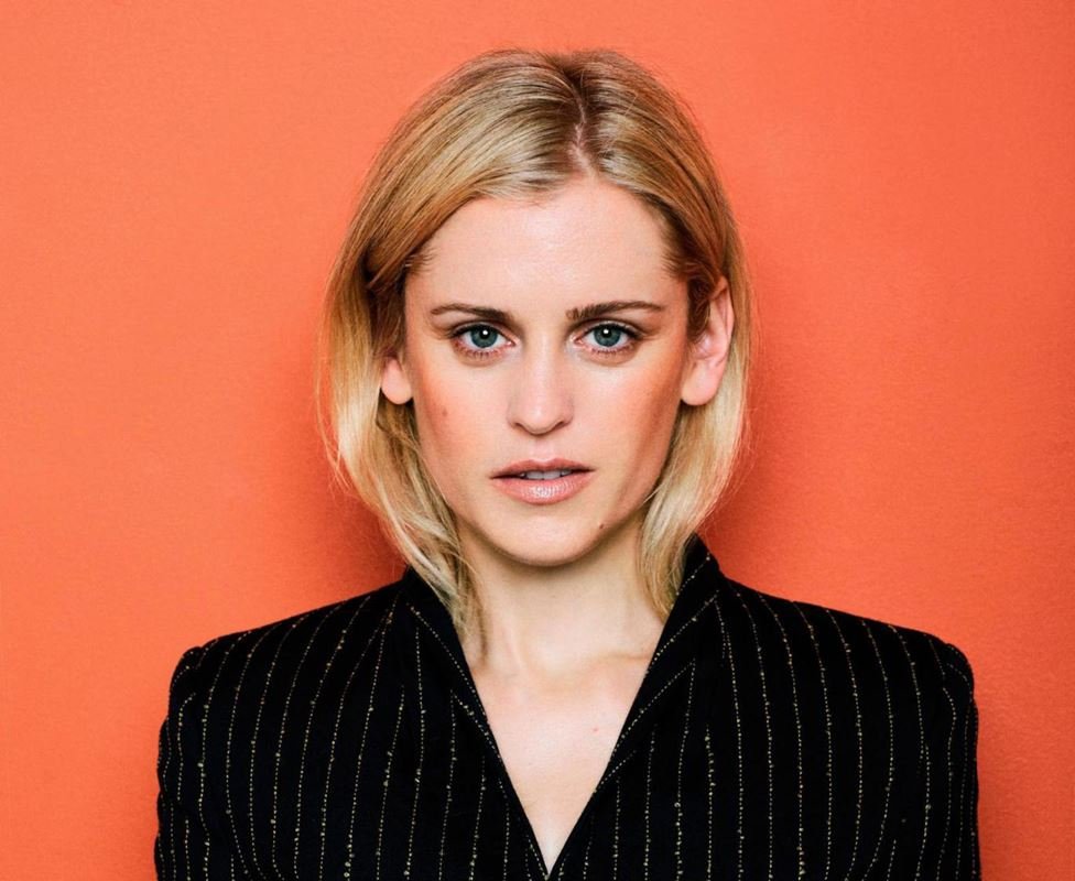 Portrait of a blonde woman with fair skin wearing a pinstripe suit jacket against an orange background in a studio headshot.