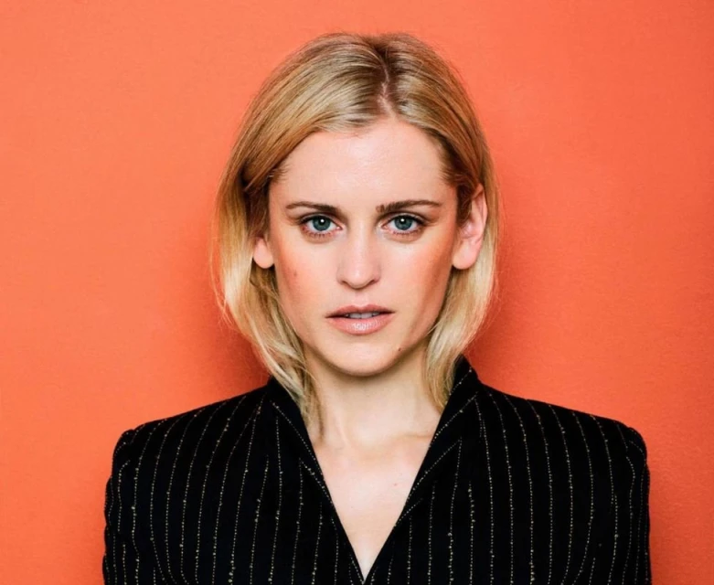 Portrait of a blonde woman with fair skin wearing a pinstripe suit jacket against an orange background in a studio headshot.