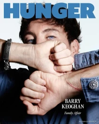 Portrait of Barry Keoghan holding his fists in front of his face against a white background, wearing a blue denim shirt.
