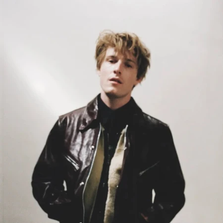 Young man with tousled blonde hair wearing a dark leather jacket against a light neutral background