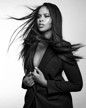 Black and white portrait of a woman with long dark hair blowing in the wind, wearing a tailored black blazer.