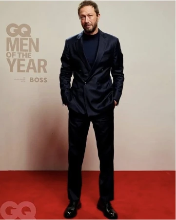 Full-length portrait of Jeremy Allen White wearing a dark blue suit standing on a red carpet against a light background