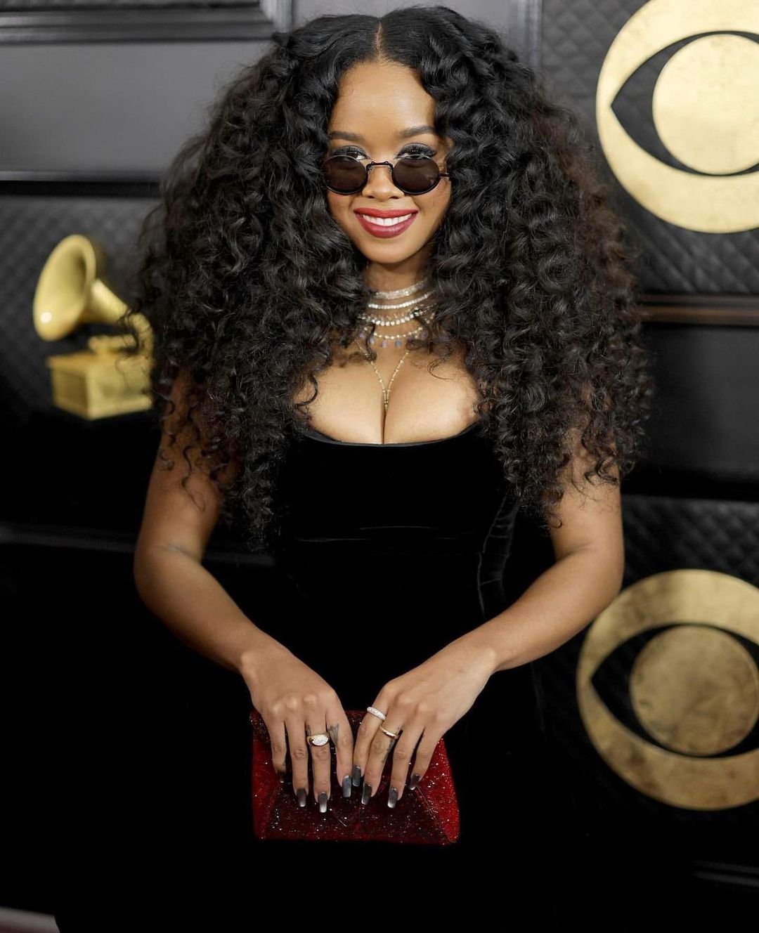 H.E.R. at Grammys: Singer with big curly hair, sunglasses, black dress, and red clutch poses at the awards show.