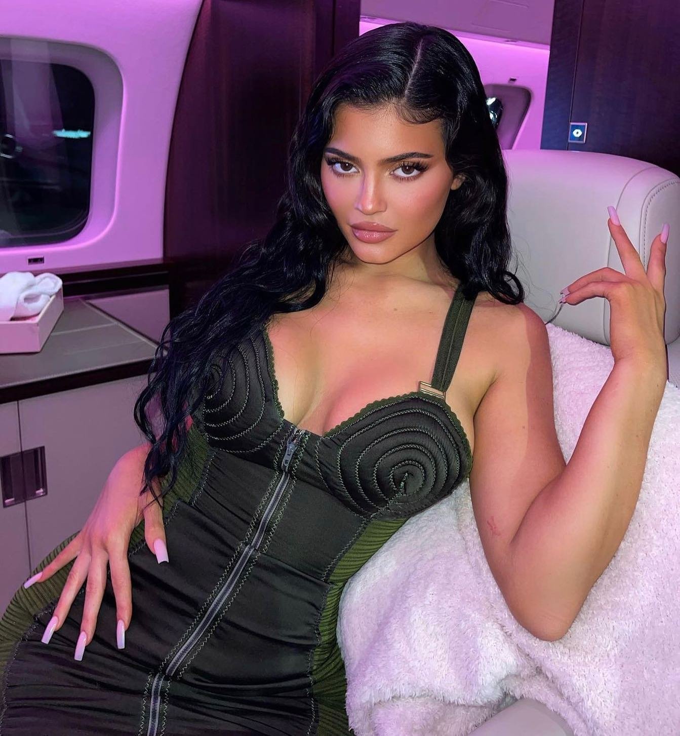 Portrait of a woman with long dark hair wearing a green dress and posing in an airplane cabin with pink interior lighting.