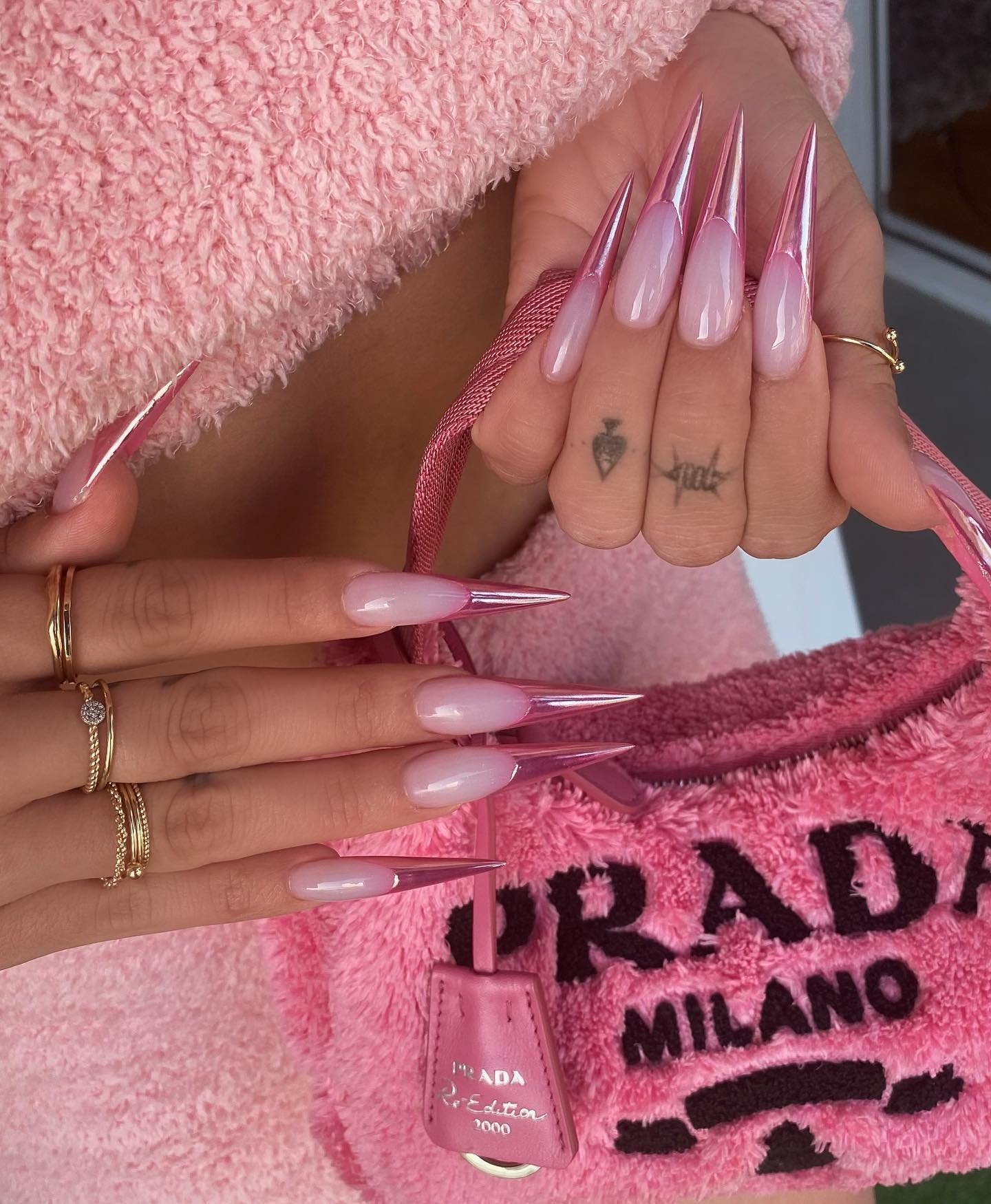Close-up shot featuring hands with long, stiletto-shaped pink and silver nails holding a pink, fuzzy Prada handbag with gold rings.