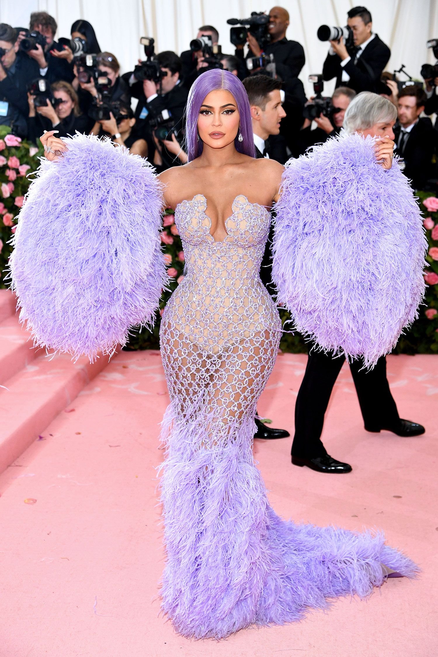 Kylie Jenner in an elaborate lavender feather dress on a pink carpet, surrounded by photographers at a celebrity event.
