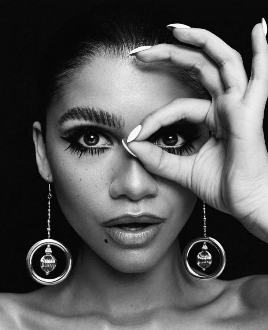 Black and white portrait of a woman with a mole on her face, circular earrings, and dark eye makeup making an okay sign.