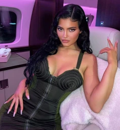 Kylie Jenner seated inside a private jet cabin wearing a dark olive green zipped dress with long dark hair and light pink manicured nails.