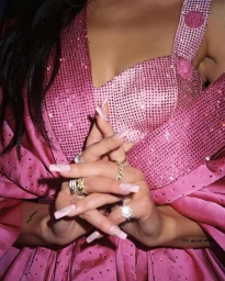 Close-up of hands with pink coffin-shaped nails wearing rings, posed against a glittering pink rhinestone dress.