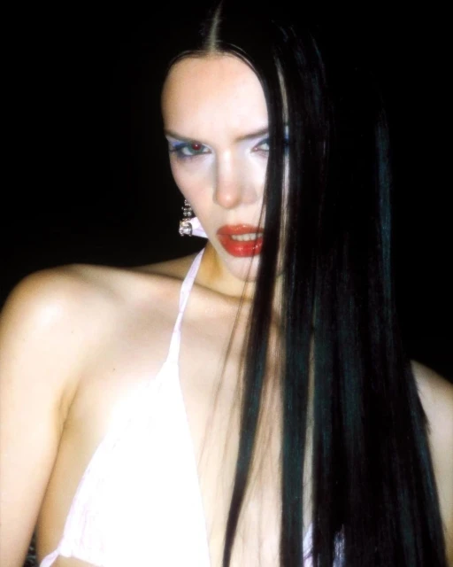 Close-up portrait of a woman with long black hair, bright red lipstick, and blue eye makeup posing against a dark black background.