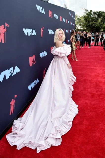 Woman in off-shoulder pink gown on a red carpet with a train, against an MTV backdrop, surrounded by blurred people and greenery.