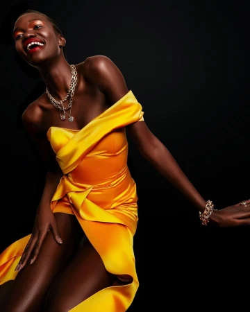 Black woman in a vibrant yellow satin off-the-shoulder dress laughing against a dark seamless background styled by Chloe Griffin.