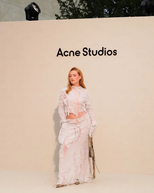 Adult female model in pale shredded-fabric long-sleeved top and maxi skirt posing against neutral background with Acne Studios text and studio light.