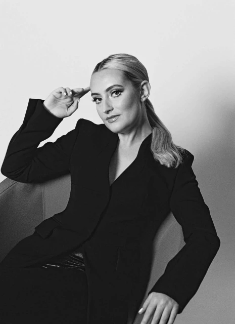 Black and white portrait of a woman with blonde hair pulled back, wearing a tailored black suit, seated against a plain backdrop.