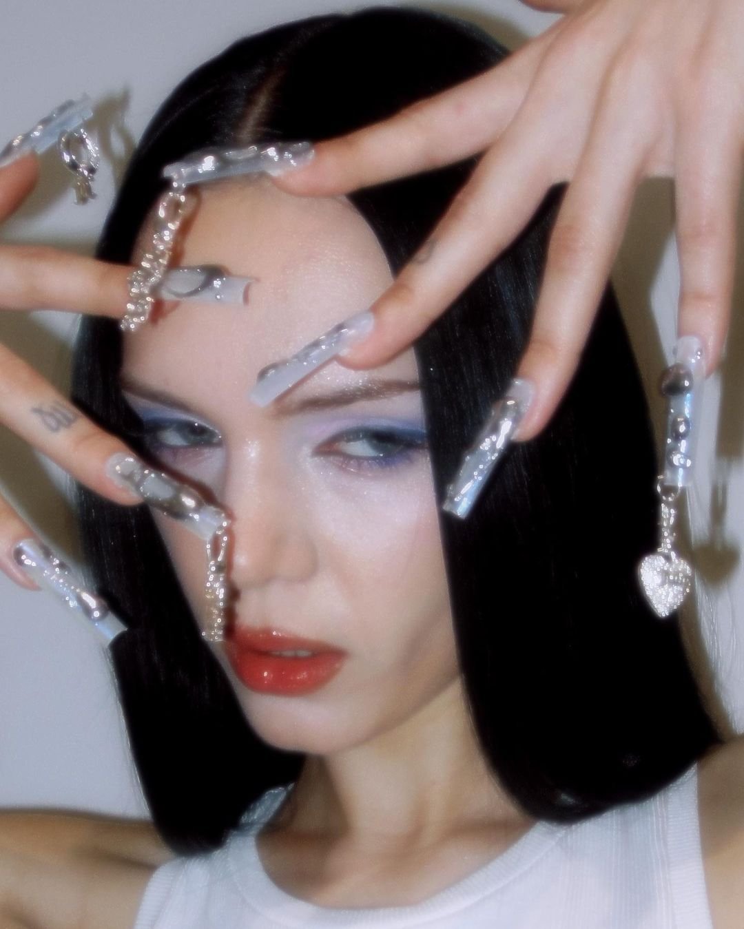 Close-up of a young woman with long, dark hair and fair skin, showcasing dramatic blue eye makeup and long, decorated nails.