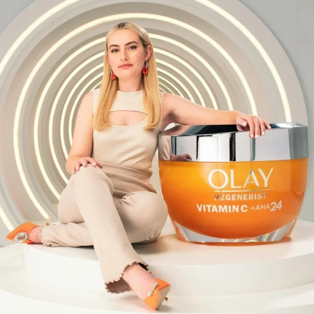 A woman in tan clothes sits beside a large container of Olay Regenerist Vitamin C + AHA 24 cream against a neutral background with circular lines.