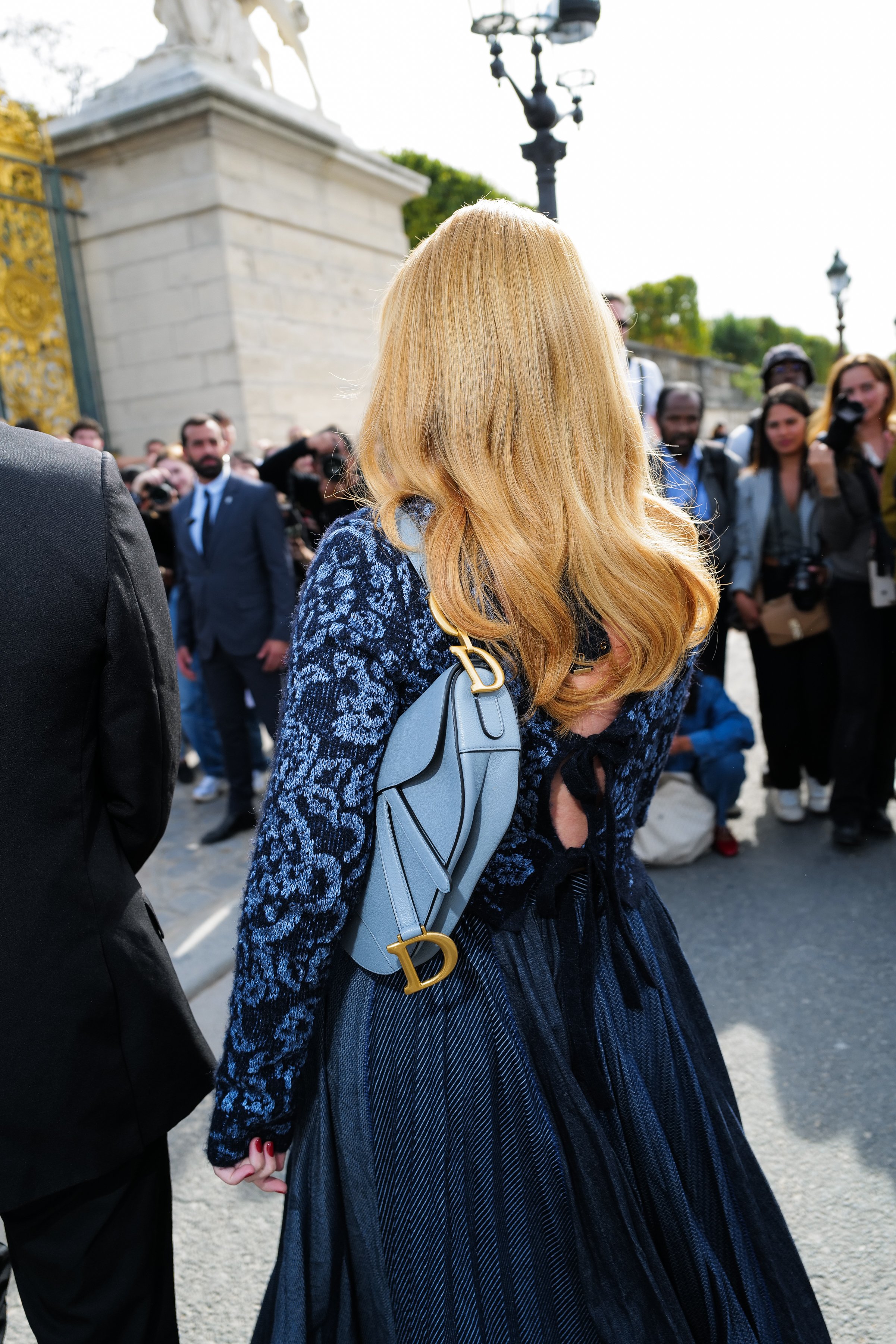 Woman with blonde hair wearing a blue dress and carrying a Dior bag, standing outdoors with a crowd in the background.