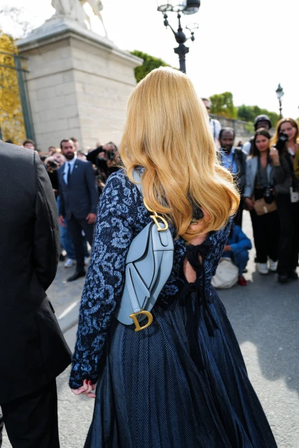 Woman with blonde hair wearing a blue dress and carrying a Dior bag, standing outdoors with a crowd in the background.