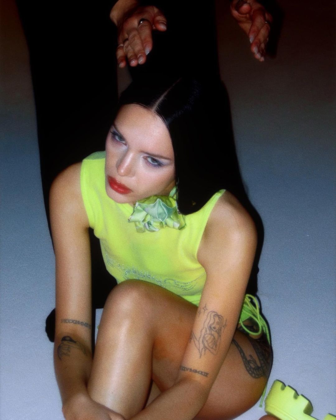 Young woman with dark hair and tattoos posing in a neon yellow top and matching platform shoes, sitting indoors with dramatic lighting.
